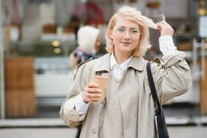 hermosa elegante mujer teniendo bueno Moda ropa caminando en calle y Bebiendo algunos café en taza para llevar con bueno verano estado animico cerca calle cafetería. foto