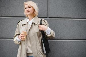 hermosa elegante mujer teniendo bueno Moda ropa caminando en calle y Bebiendo algunos café en taza para llevar con bueno verano estado animico cerca calle cafetería. foto