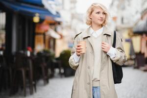 hermosa elegante mujer teniendo bueno Moda ropa caminando en calle y Bebiendo algunos café en taza para llevar con bueno verano estado animico cerca calle cafetería. foto
