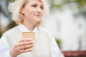 hermosa elegante mujer teniendo bueno Moda ropa caminando en calle y Bebiendo algunos café en taza para llevar con bueno verano estado animico cerca calle cafetería. foto