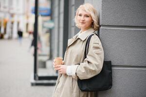 hermosa elegante mujer teniendo bueno Moda ropa caminando en calle y Bebiendo algunos café en taza para llevar con bueno verano estado animico cerca calle cafetería. foto