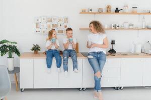 Mom with her two children sitting on the kitchen table. Mother with daughter and toddler son having breakfast at home. Happy lifestyle family moments. photo