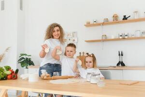 contento joven madre con linda pequeño preescolar niños tener divertido haciendo masa horneando tarta o Pastelería en moderno cocina juntos, lleno de alegría madre enseñar pequeño niños haciendo panadería Cocinando a hogar foto