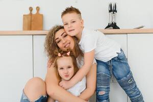 Happy mother hugging daughter and son sitting on wooden floor in modern kitchen at home. Mom hugging children. Happy family relationship concept photo