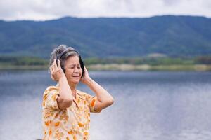 Portrait of a happy elderly Asian woman wearing wireless headphones listening to music for relaxing standing at the side of the lake. Space for text. Concept of aged people and relaxation photo