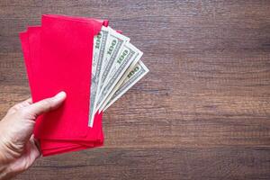 Close-up of hand holding a red envelope packet with money dollars on wooden table background. Space for text. Concept of money photo