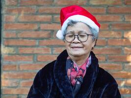 Portrait of an elderly Asian woman wearing a red hat with eyes closed and smiling while standing with brick wall background photo