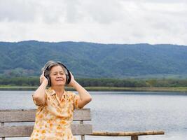 Portrait of senior Asian woman wearing wireless headphones listening to a favorite song while sitting on a wooden chair side the pond. Concept of aged people and relaxation photo