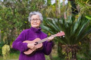 Portrait of an elderly Asian woman with short gray hair playing the ukulele while standing in a garden. Concept of aged people and relaxation photo