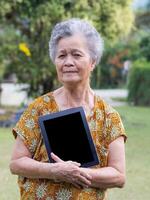 Senior woman with short gray hair holding a tablet and looking at the camera while standing in a garden. Space for text. Concept of aged people and technology photo