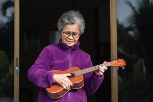 alegre mayor asiático mujer con corto gris pelo vistiendo lentes y jugando el ukelele mientras en pie en frente de el puerta. concepto de Envejecido personas y relajación foto