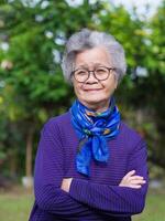 retrato de un mayor mujer con corto gris cabello, sonriente, brazos cruzado, y mirando a el cámara mientras en pie en un jardín. concepto de Envejecido personas y cuidado de la salud foto