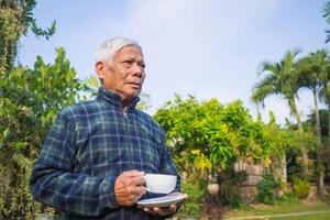 Portrait of a senior man holding a white cup of coffee while standing in a garden. Space for text. Concept of aged people and relaxation photo