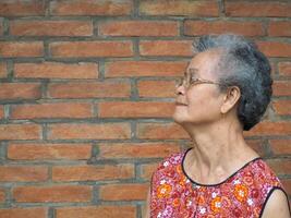 lado ver de un mayor mujer sonriente y mirando lejos mientras en pie con ladrillo pared antecedentes. espacio para texto. concepto de personas mayores y cuidado de la salud foto