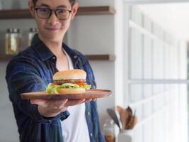 Close-up of a homemade hamburger on a wooden dish, holding by young man stand wearing glasses looking at the camera, smiling with blur background. Unhealthy Foods concept. Junk-food photo