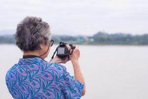 mayor mujer con corto gris pelo vistiendo Gafas de sol, tomando un foto por un digital cámara a el orilla con montañas antecedentes. concepto de Envejecido personas y fotografía