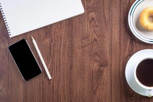 Top view of a smartphone, sketchbook, a white coffee cup, and donut on a plate placed on a wooden table. Template for text. Wood texture background. Concept of drawing and technology photo