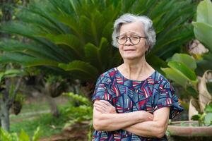 A portrait of an elderly woman with short gray hair wearing glasses, smiling with arms crossed, and looking at the camera while standing in a garden. Concept of aged people and healthcare photo