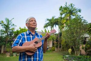 retrato de un contento mayor hombre jugando el ukelele, sonriente y mirando arriba mientras en pie en un jardín. espacio para texto. concepto de Envejecido personas y relajación foto