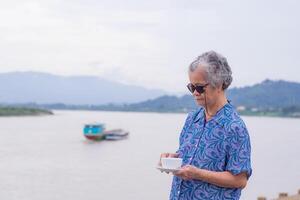 Senior woman with short gray hair wearing sunglasses and holding a white coffee cup while standing beside the river with a nature background. Morning coffee. Space of text photo