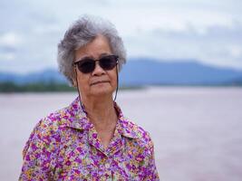 Portrait of elderly Asian woman wearing sunglasses and looking at the camera while standing beside the river. Space for text. Concept of aged people and relaxation photo