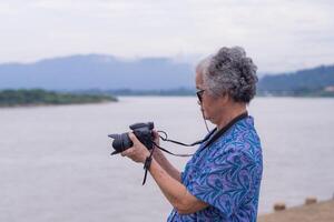 un mayor mujer con corto gris pelo participación un digital cámara mientras en pie lado el río con naturaleza antecedentes. Mañana café. concepto de Envejecido personas y fotografía foto