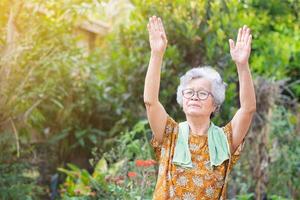 retrato de mayor asiático mujer ejercicio por gesticulando aumento ambos brazos mientras en pie en un jardín. un hermosa mayor mujer es contento y saludable. concepto de Envejecido personas y cuidado de la salud foto