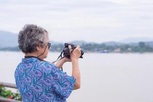 mayor mujer con corto gris pelo vistiendo Gafas de sol, tomando un foto por un digital cámara a el orilla con montañas antecedentes. concepto de Envejecido personas y fotografía