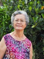 retrato de un mayor mujer con corto gris cabello, vistiendo anteojos, sonriente y mirando a el cámara mientras en pie en un jardín. espacio para texto. concepto de Envejecido personas y cuidado de la salud foto