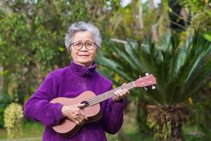 alegre mayor asiático mujer con corto gris pelo vistiendo lentes y jugando el ukelele mientras en pie en un jardín. concepto de Envejecido personas y relajación foto