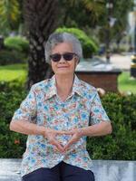 Portrait of a happy elderly Asian woman with short gray hair, wearing sunglasses, smiling and showing fingers heart shape symbol while sitting on a chair in the park. Aged people concept photo