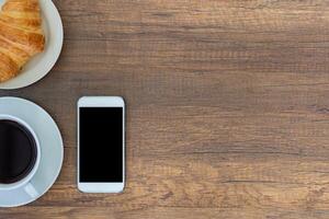 Top view of a table with a smartphone, a white coffee cup, and croissants placed on a wooden table. Work from home. Space for text photo