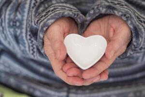 de cerca de un en forma de corazon blanco cerámico en el manos de un mayor mujer. concepto de Envejecido personas y cuidado de la salud foto