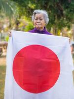 Portrait of elderly woman with short white hair holding Japanese flag, standing in her garden. photo