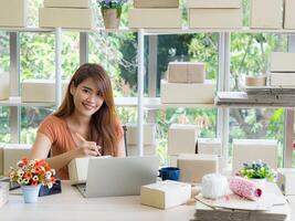 Businesswoman smiling and write data on cover parcel box for prepare send to a customer while sitting on a chair in the office. Concept of business and e-commerce photo