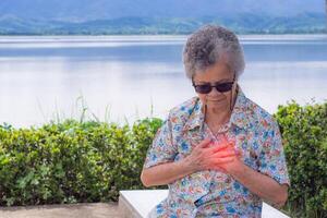 un mayor mujer agarrando su cofre en dolor a el primero señales de angina de pecho o miocárdico infarto o corazón ataque mientras sentado lado lago. concepto de Envejecido personas y corazón ataque foto