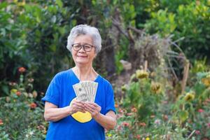 Portrait of a senior woman with short gray hair, hands holding US banknotes, and looking at the camera while standing in a garden. Selective focus. Concept of aged people and finance photo