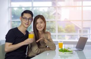 asiático joven Pareja participación un vaso de Fresco naranja jugo para salud con vegetal ensalada, computadora portátil, y un teléfono inteligente metido en el mesa mientras sentado en el cocina. concepto de amor Pareja foto