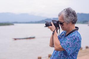 Senior woman with short gray hair wearing sunglasses, taking a photo by a digital camera at the riverside with mountains backgrounds. Concept of aged people and photography