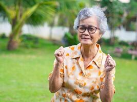 Portrait of happy senior woman clenching fists excited for success with arms raised celebrating victory smiling while standing in a garden. Concept of aged people and healthcare photo