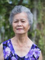 retrato de un mayor mujer con corto gris pelo en pie sonriente y mirando a el cámara mientras en pie exterior. concepto de Envejecido personas y cuidado de la salud foto