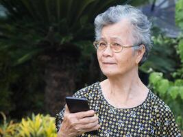 un retrato de un mayor mujer utilizando un teléfono inteligente y mirando lejos mientras en pie en un jardín. espacio para texto. concepto de Envejecido personas y tecnología foto