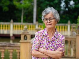 Portrait of senior Asian woman wearing glasses, arms crossed, and looking at the camera while standing in the park. Concept of aged people and healthcare photo