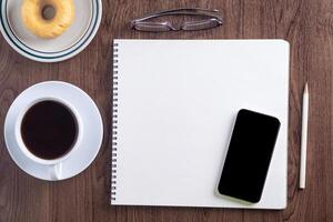 Top view of a smartphone, sketchbook, a white coffee cup, and donut on a plate placed on a wooden table. Template for text. Wood texture background. Concept of drawing and technology photo