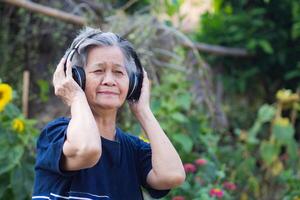 Elderly woman standing wearing wireless headphone listening to a favorite song, and smile happy in garden. Asian senior woman, short white hair are enjoying the music. Good mental health concept photo