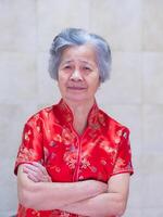 An elderly woman wearing a traditional cheongsam qipao dress, smiling and arms crossed while standing at home. Chinese woman in red costume New Year celebration. Concept of aged people Chinese photo