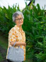 Portrait of a happy senior woman smiling, looking at the camera, and posing with a shopping bag while standing in a garden. Space for text. Concept of aged people and shopping photo