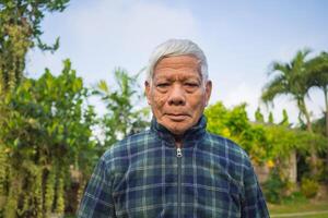 retrato de un mayor asiático hombre sonriente y mirando a el cámara mientras en pie en un jardín. espacio para texto. concepto de Envejecido personas y cuidado de la salud foto