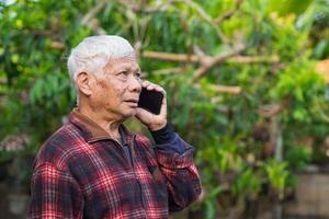 un mayor asiático hombre utilizando un teléfono inteligente y mirando arriba mientras en pie en el jardín. espacio para texto. concepto de Envejecido personas y tecnología foto