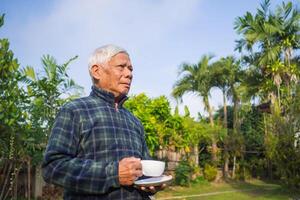 Portrait of an elderly man holding a cup of coffee and looking away while standing in a garden. Concept of aged people and relaxation photo